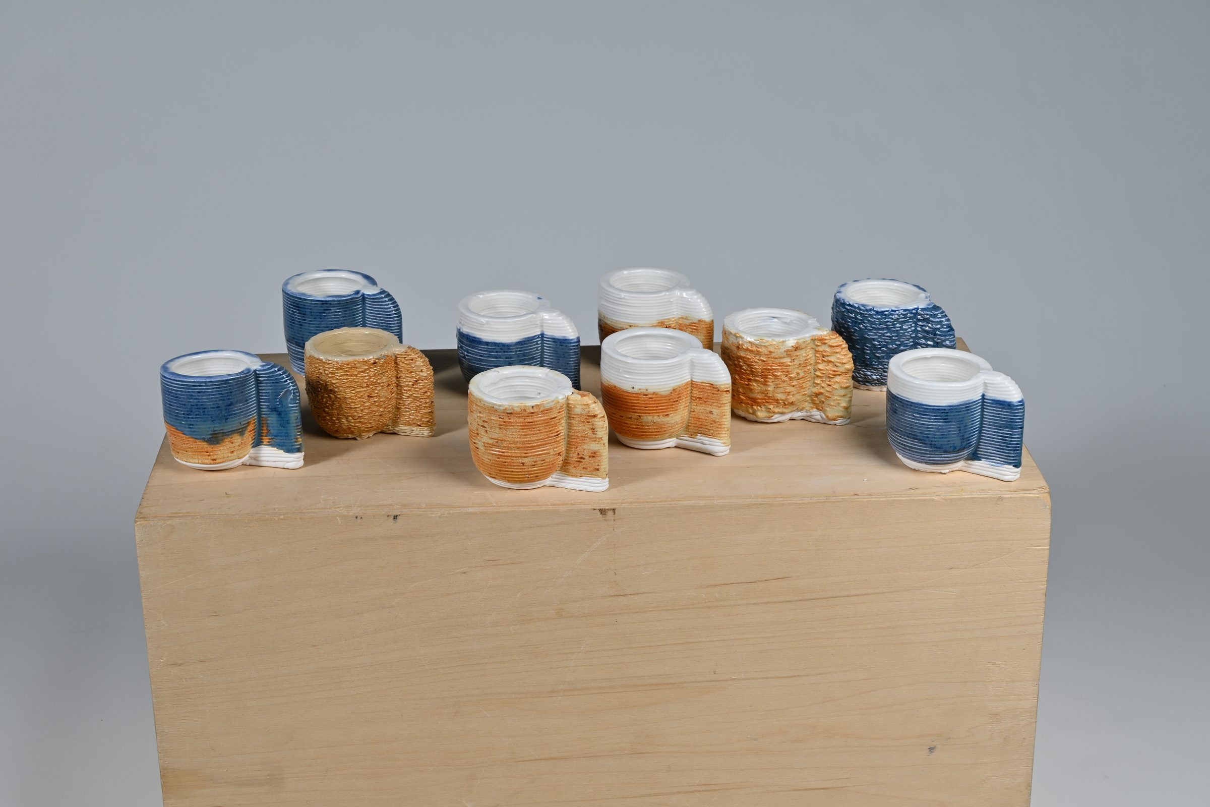 A row of printed ceramic cups in blue, white and terracotta glazes on a plinth